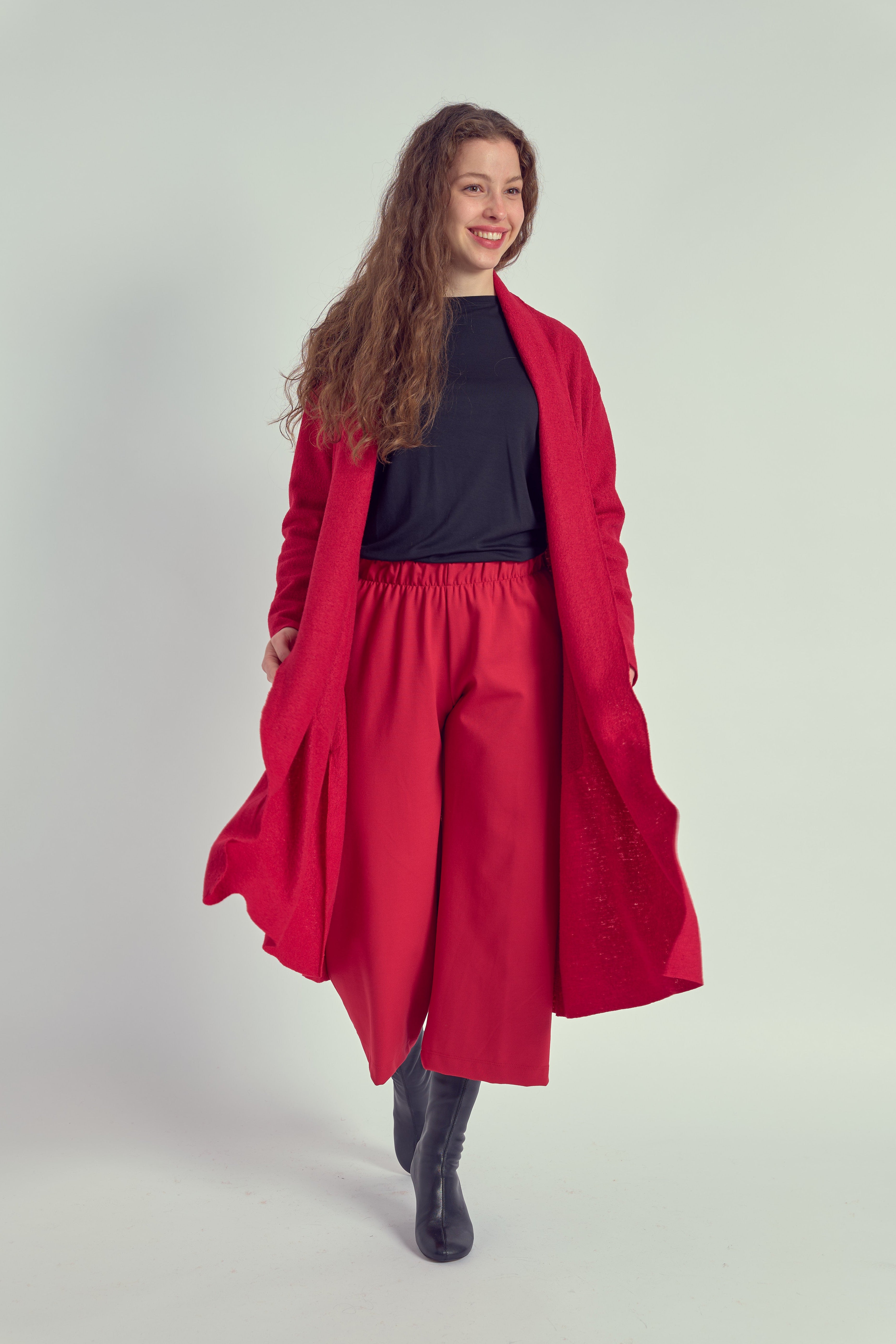 A person wearing a long red cardigan with patch pockets, paired with a dark top and red trousers, standing against a white background.