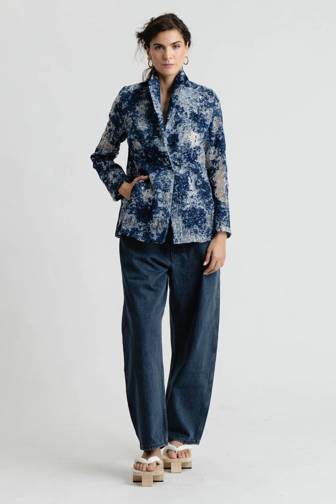 A model stands against a light gray studio background wearing a blue and white abstract patterned blazer, dark blue wide-leg jeans, and beige platform sandals.