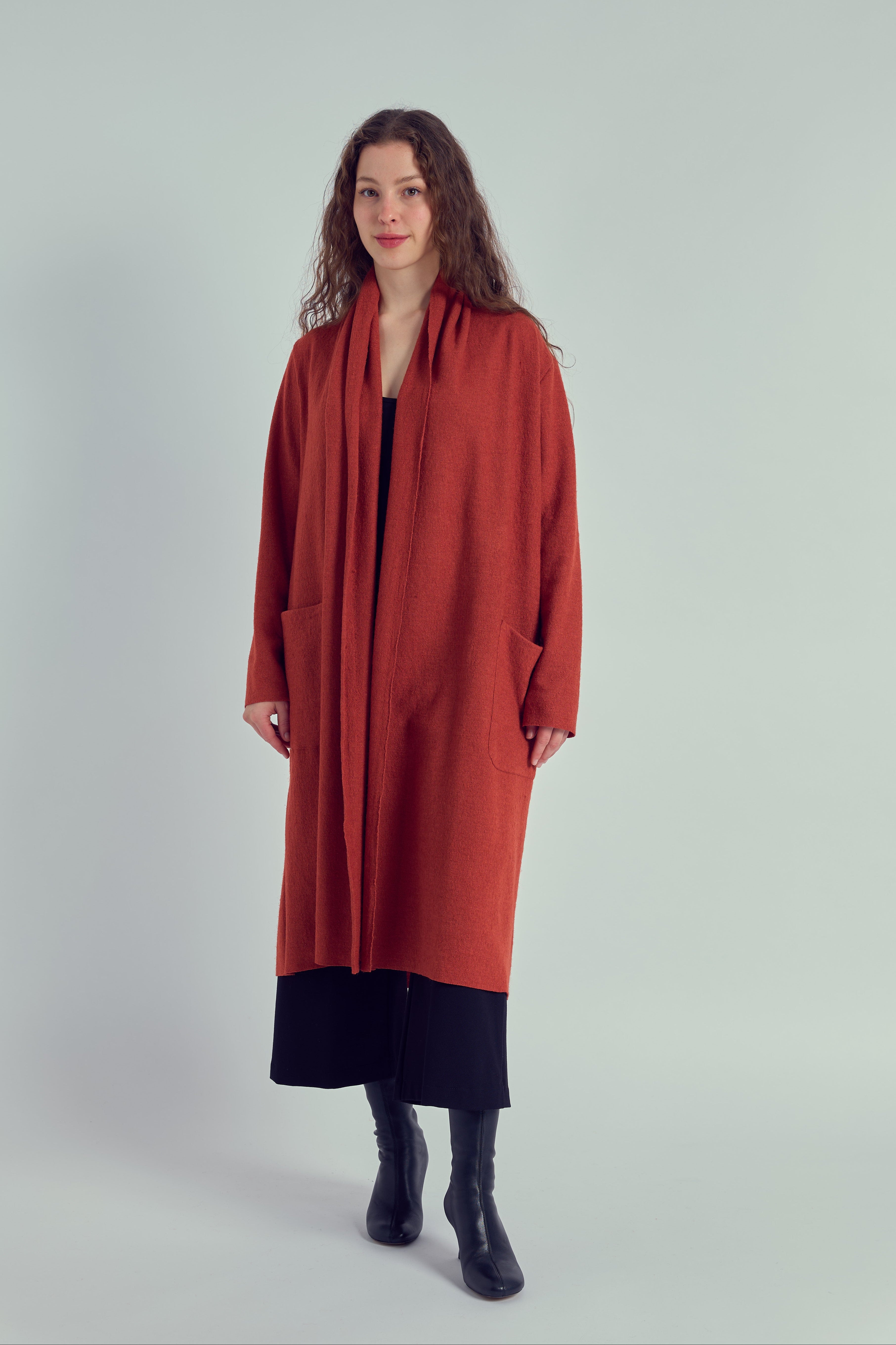 A person wearing a long rust colour cardigan with patch pockets, paired with a dark top and trousers, standing against a white background.