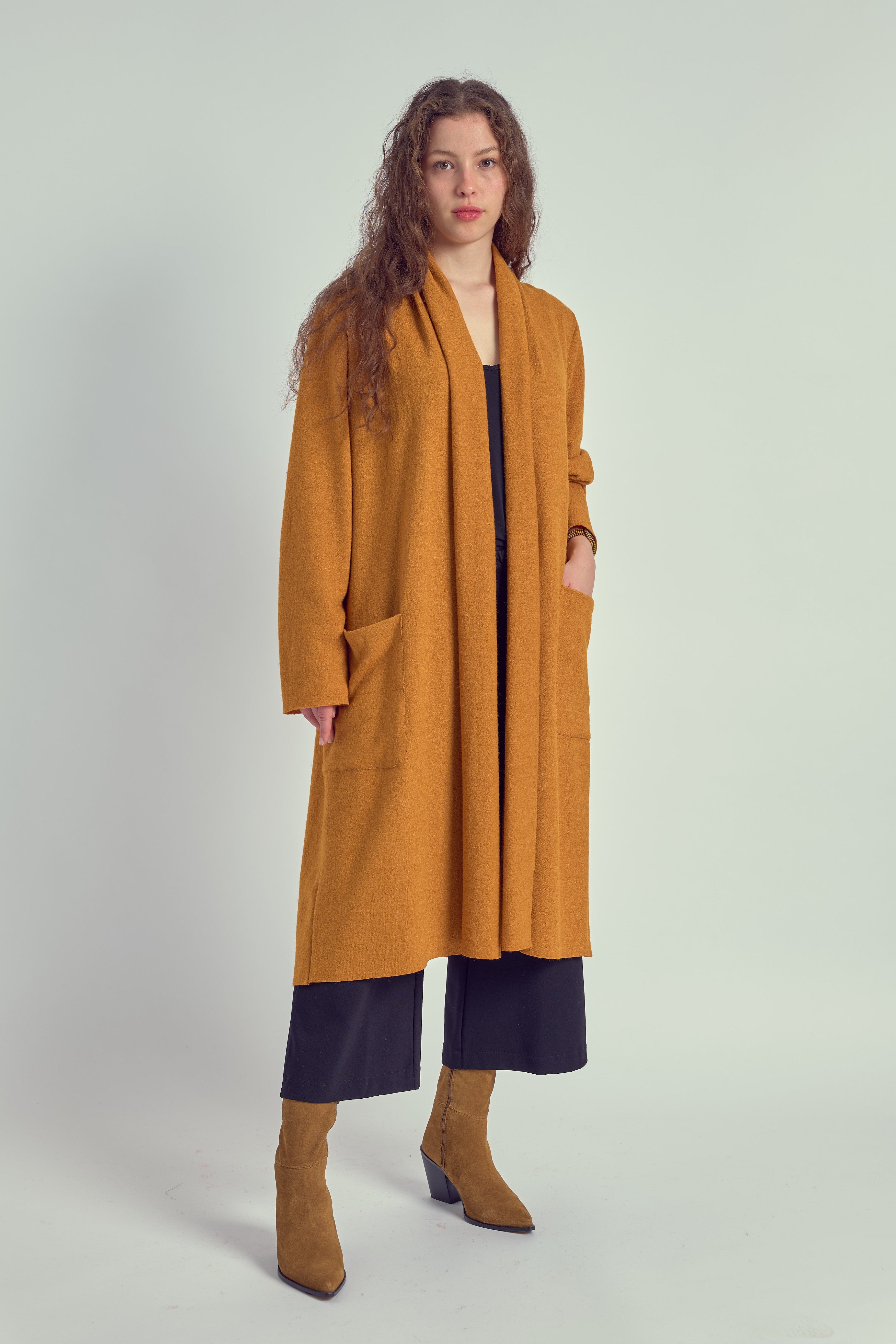 A person wearing a long purple cardigan with patch pockets, paired with a dark top and trousers, standing against a white background.