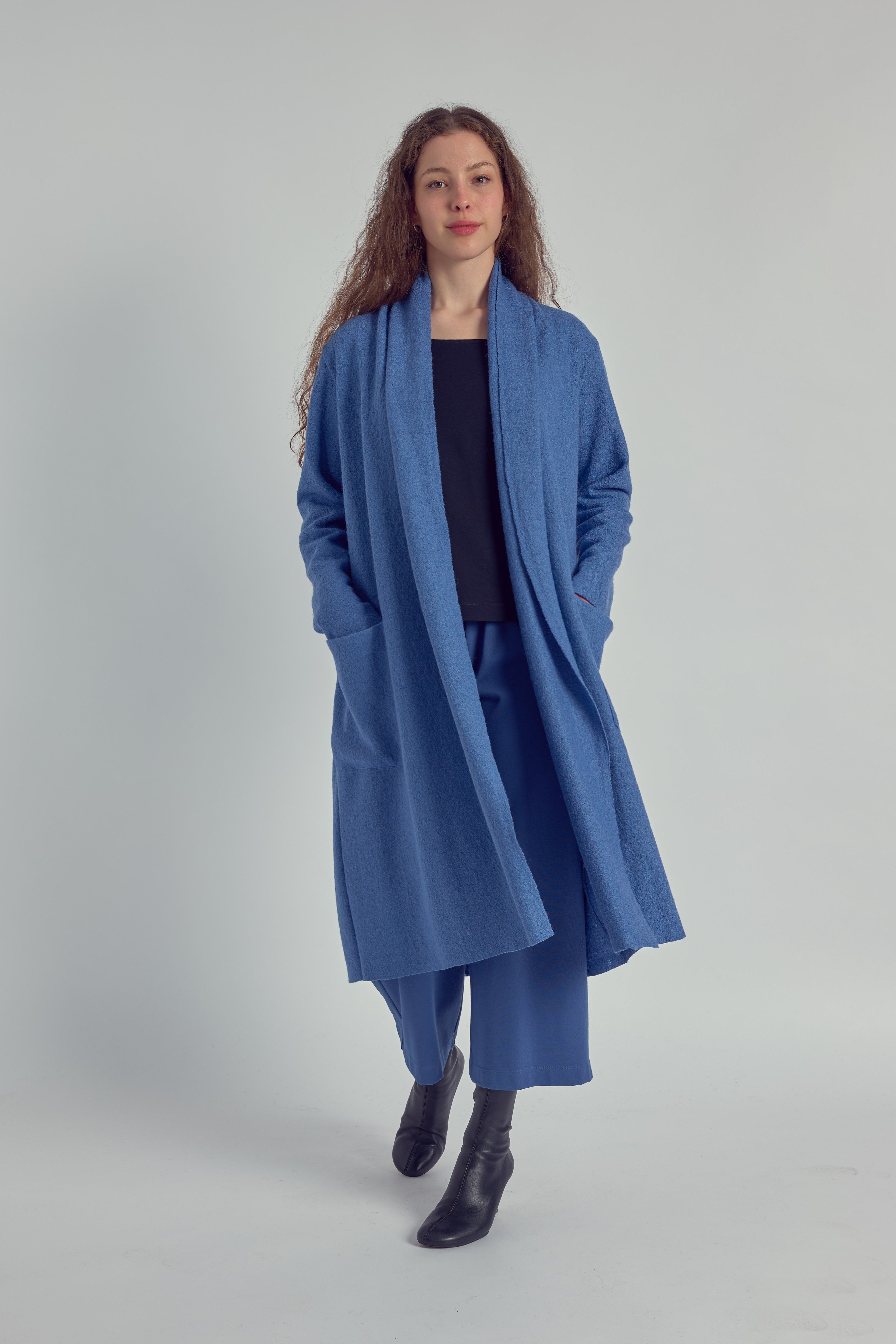 A person wearing a long sky blue cardigan with patch pockets, paired with a navy top and sky blue trousers, standing against a white background.