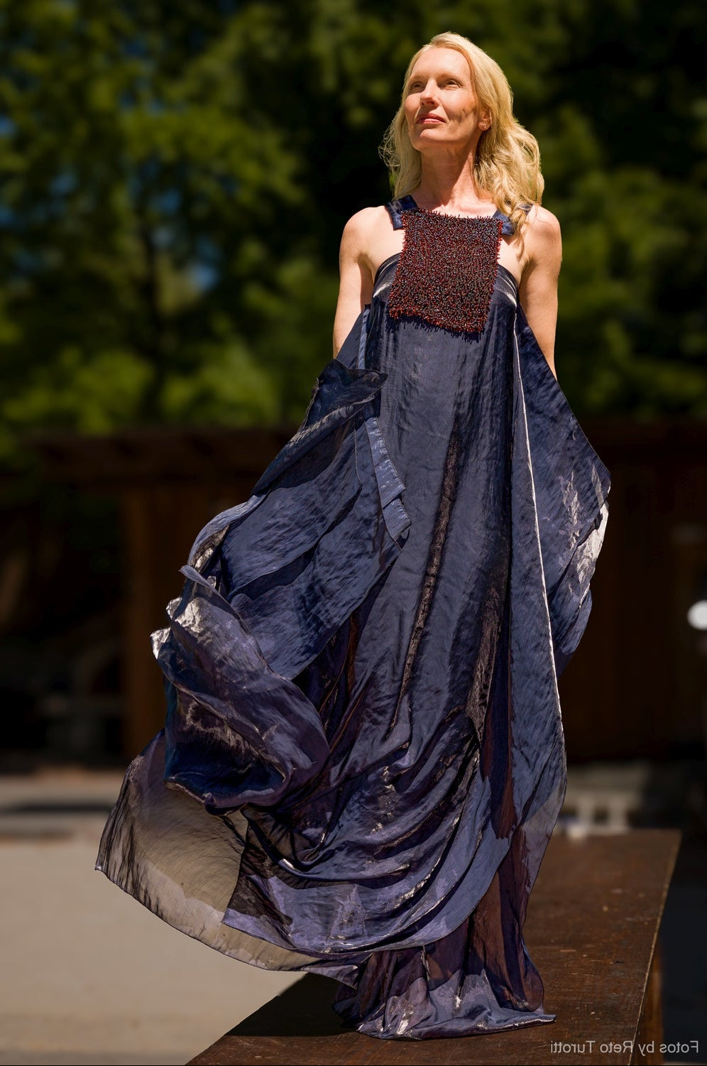 A woman wearing a flowing sleeveless maxi dress in dark navy blue and black, standing outdoors on a wooden platform with trees in the background.